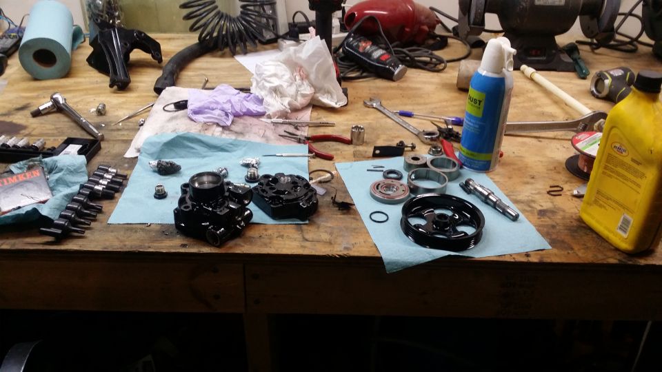 Various automotive parts, including a power steering pump and pulley, laid out on a wooden workbench with tools and cleaning supplies