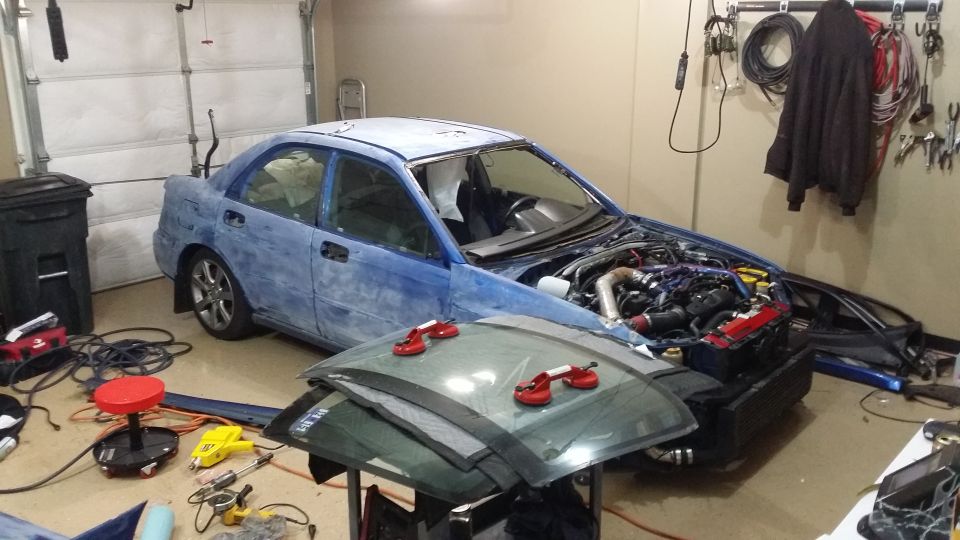 Car in garage with hood open, engine exposed. Windshield removed and stacked in front