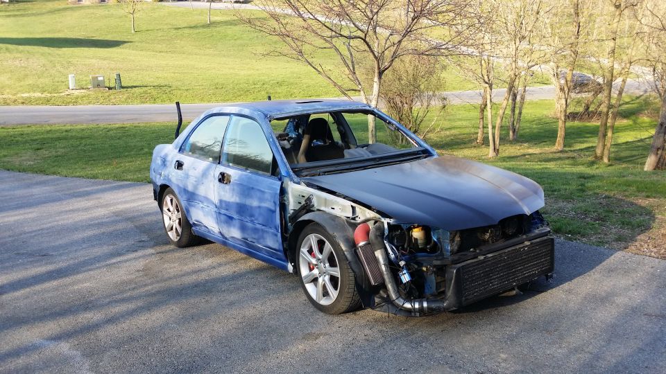 Blue Subaru Impreza missing front bumper, hood, and fenders. Aftermarket intercooler and intake piping are visible. Racing seat inside