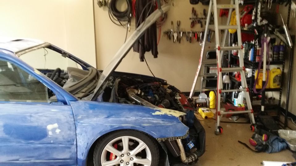 Blue Subaru Impreza with hood open, showing engine bay and front fender damage. Ladder and tools in background