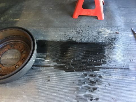 1986 Jeep Wagoneer - A brake drum sits on a ribbed floor next to a dark fluid spill, with a red jack stand visible.