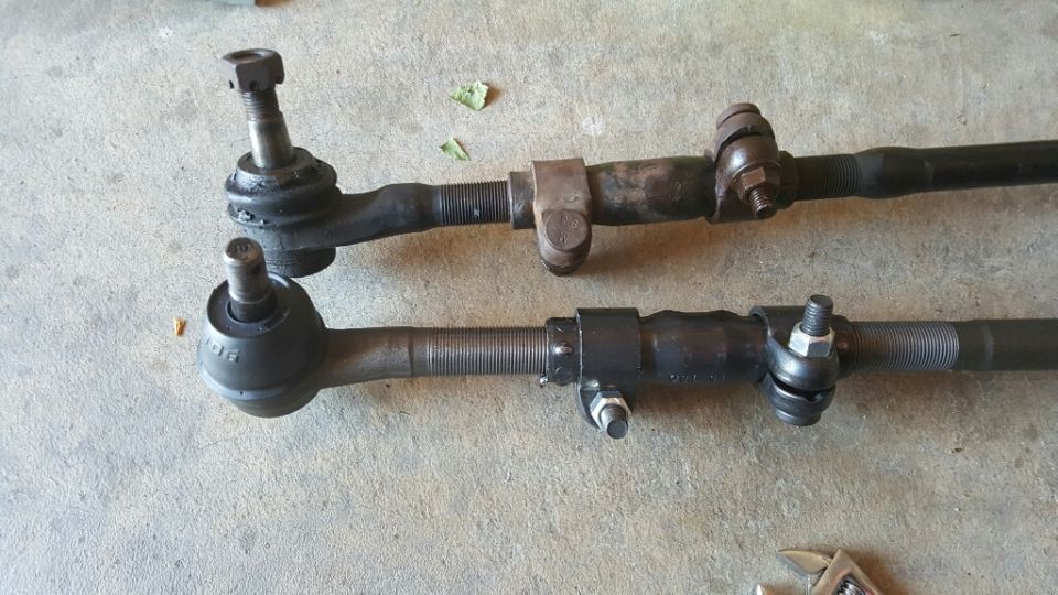 Two steering tie rods, one above the other, on a concrete surface. The bottom one has a black boot and a nut on the ball joint