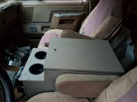1988 Ford Bronco - The interior of a vehicle shows tan fabric seats and an aftermarket center console with cup holders.