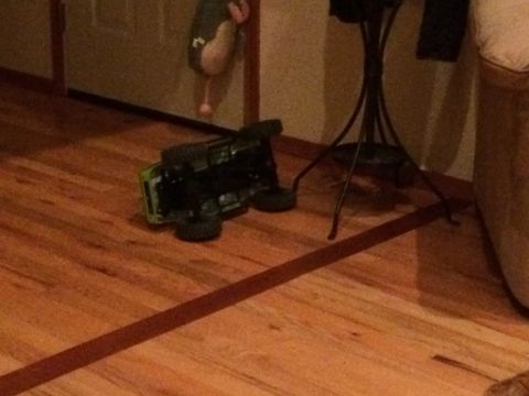 1989 Ford Bronco - A green toy truck lies overturned on a polished wooden floor, with a decorative stand and part of a couch visible in the background.