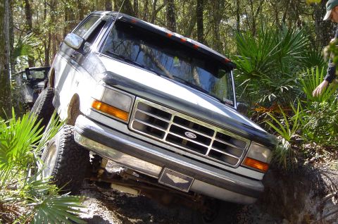 1993 Ford Bronco - A white and black Ford Bronco navigates a challenging off-road trail, leaning heavily to one side.