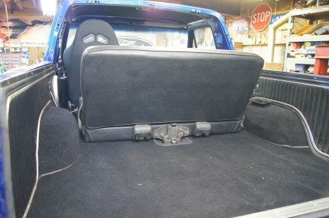 1979 Ford Bronco - The interior rear cargo area of a blue Ford Bronco is shown, featuring a black bench seat folded up against the rear wall and black carpeti…