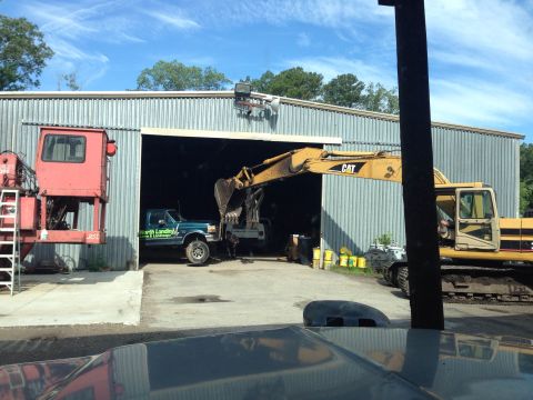 1979 Ford Bronco - A yellow CAT excavator is parked outside a large metal building, with its arm extended into an open bay where a teal Ford pickup truck is p…