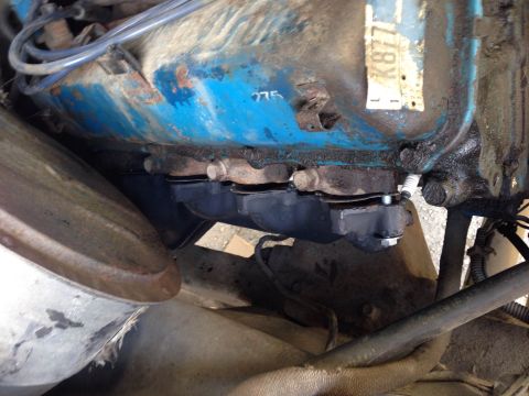 1979 Ford Bronco - A close-up view of the exhaust manifold and part of the engine block of a vehicle. The engine block is painted blue and shows signs of rust…