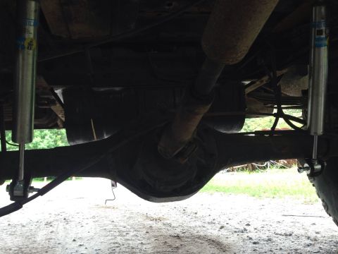 1979 Ford Bronco - Underside view of a vehicle's rear axle and suspension system, featuring two silver shock absorbers with blue and yellow branding. A portio…