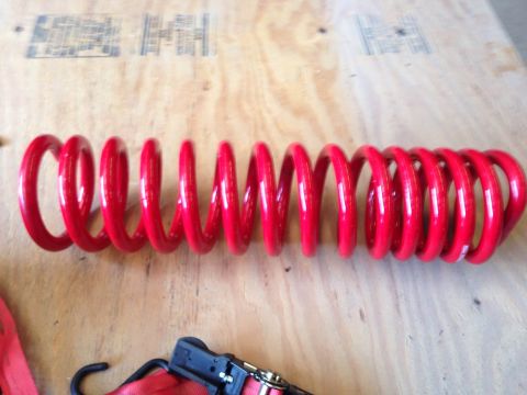 1979 Ford Bronco - A bright red coil spring is laid horizontally on a light brown wooden surface. The spring is the primary focus, with some red straps and a…