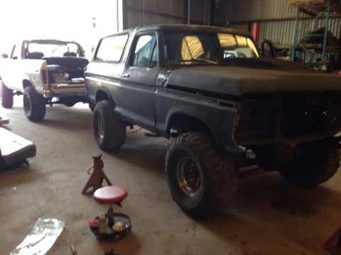 1979 Ford Bronco - A dark-colored Ford Bronco, missing its front grille and headlights, is parked indoors next to a white pickup truck. The Bronco has large o…