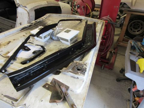 Car parts laid out on a workbench, some black, some metal. A car body is visible in the background