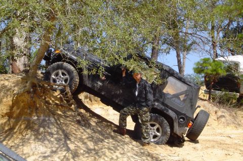 1993 Ford Bronco - A person stands by a Jeep Wrangler as it navigates a steep, sandy incline surrounded by trees.