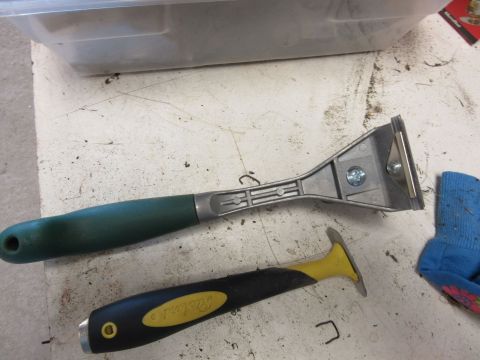 Two razor blade scrapers, one with a green handle and one with a yellow and black handle, sit on a dirty surfa