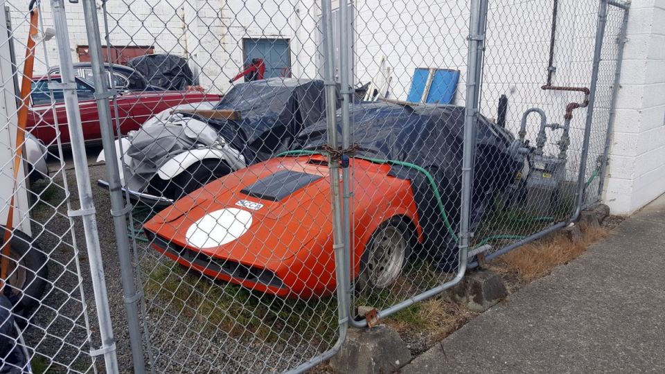 Orange sports car with white decal on hood, behind fence, with red classic car behind it.