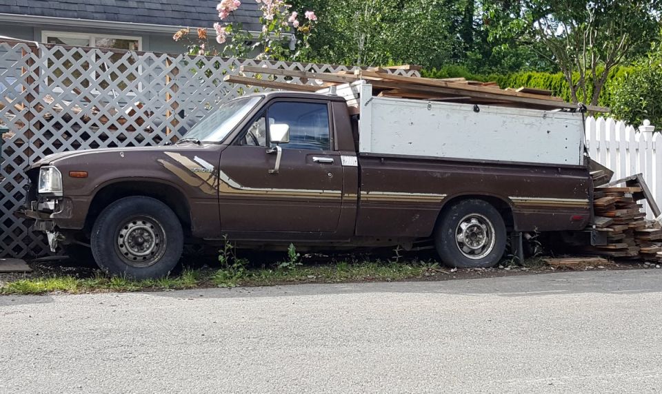 Brown pickup truck with white utility cap loaded with lumber, parked on the side of the road.