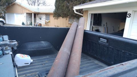 1978 Ford Bronco - The interior of a truck bed holds two large pipes and a white jug, with a house and garage in the background.