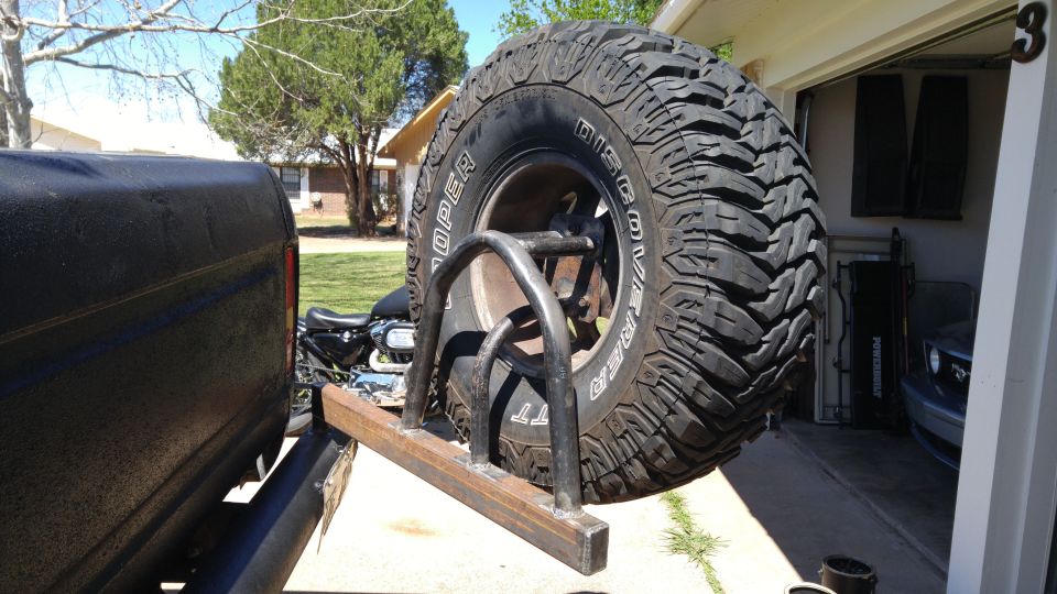 Spare tire mounted on a custom rack on the back of a dark-colored vehicle. A motorcycle is visible behind it. A garage with a car is in the background