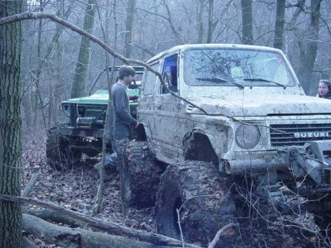 1992 Ford Bronco - Two off-road vehicles are parked in a wooded area with large tires and muddy bodies.