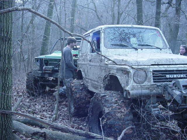 Two off-road vehicles in a wooded area, one is a white Suzuki Samurai with large tires