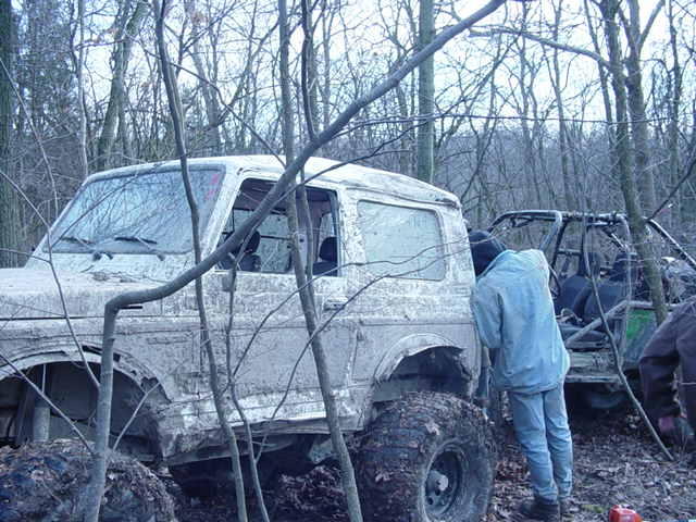 Muddy off-road vehicles parked in a wooded area with bare trees. One vehicle is a white SUV, and another is a green ATV