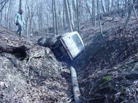 1992 Ford Bronco - A white SUV is stuck on its side in a steep, wooded ravine, with a person observing from the embankment.