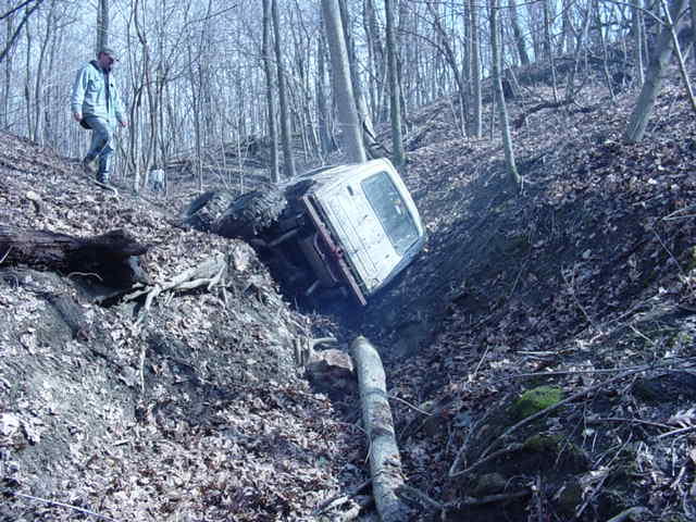 White SUV tipped on its side in a wooded ditch, with a person standing on the bank above