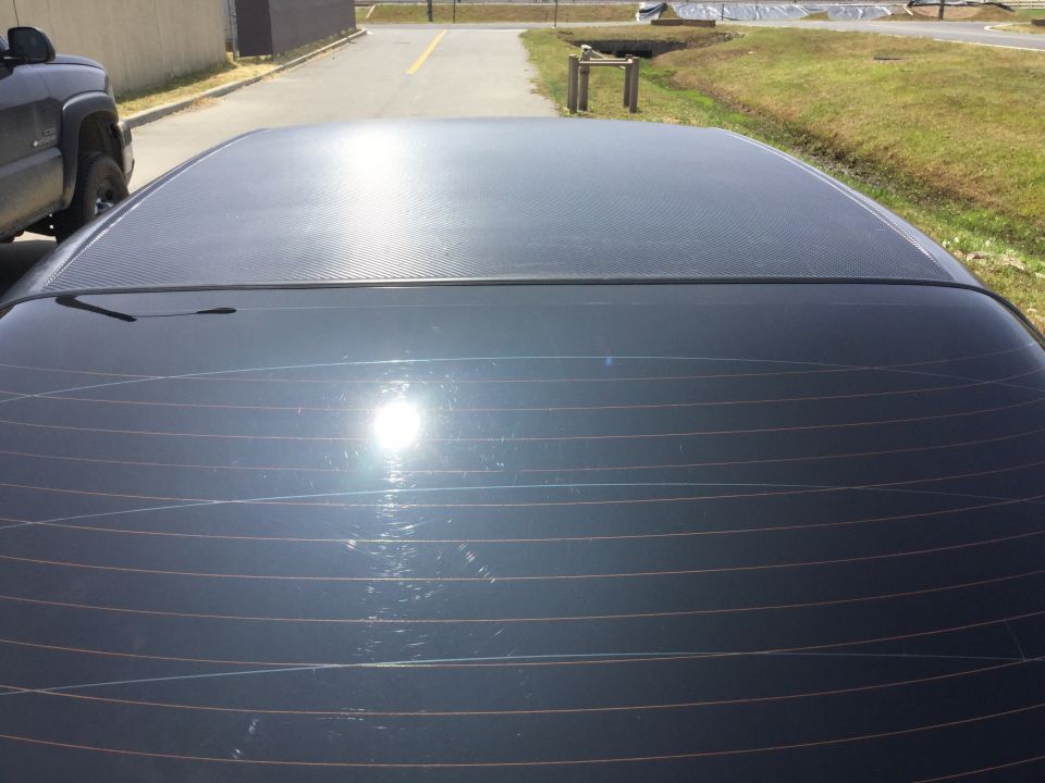 Dark colored car roof and rear window with defroster lines. A black pickup truck is parked to the left