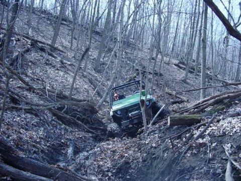 1992 Ford Bronco - A green off-road vehicle makes its way down a challenging, debris-strewn slope in a dense forest.