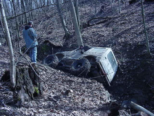 Off-road vehicle tilted in a muddy ditch with a person standing nearby on a wooded hillside