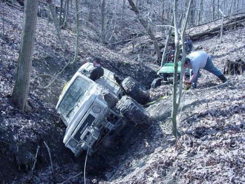 1992 Ford Bronco - Two off-road vehicles navigate a steep, wooded terrain with one vehicle partially submerged in a muddy ditch.