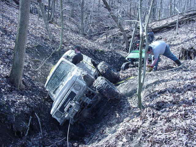 White Suzuki Samurai stuck in a ditch with large tires, a person in the driver's seat, and another person working nearby