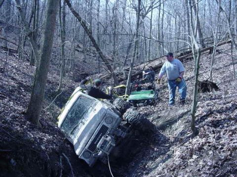 1992 Ford Bronco - A white SUV is stuck in a ditch in a wooded area, with a green ATV and people nearby.