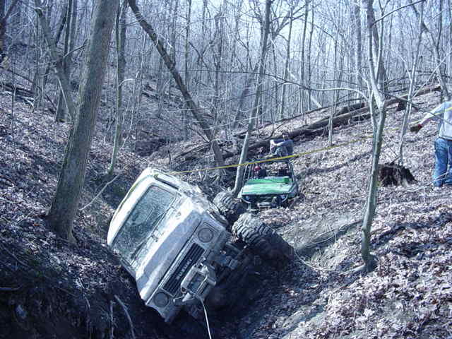 Two off-road vehicles, one white SUV and one green ATV, stuck in a wooded ditch with people nearby
