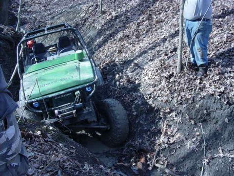 1992 Ford Bronco - An off-road vehicle navigates a muddy, wooded terrain, with the Suzuki logo clearly visible on the front.