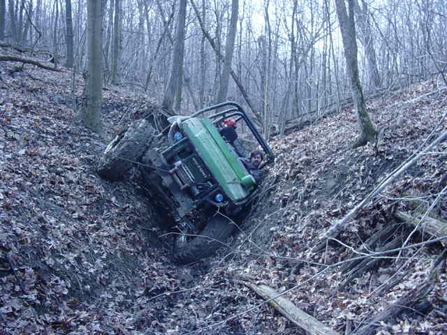 Green off-road vehicle tilted on a steep, muddy, wooded slope with a person inside