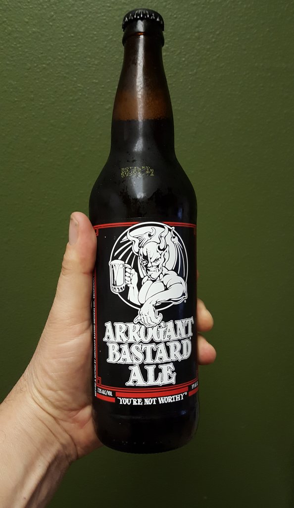 Hand holding a dark glass beer bottle with a black label a devil graphic and "ARROGANT BASTARD ALE" text