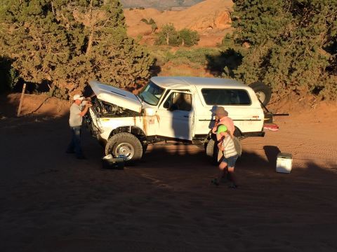 1979 Ford Bronco - Two people are working on the engine of a white SUV in a desert environment while another person and a child observe.