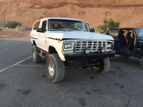 1979 Ford Bronco - A modified white Ford Bronco sits parked on asphalt, with a rocky cliff face in the background and another off-road vehicle to its right.