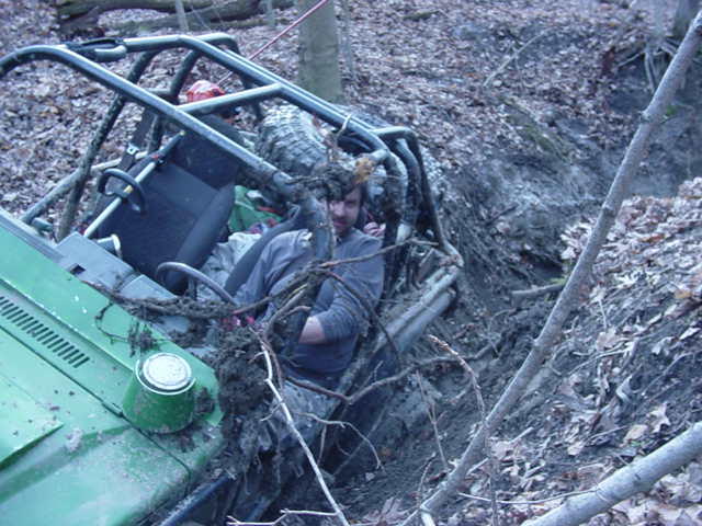 Green off-road vehicle with roll cage stuck in a muddy ditch, driver visible inside