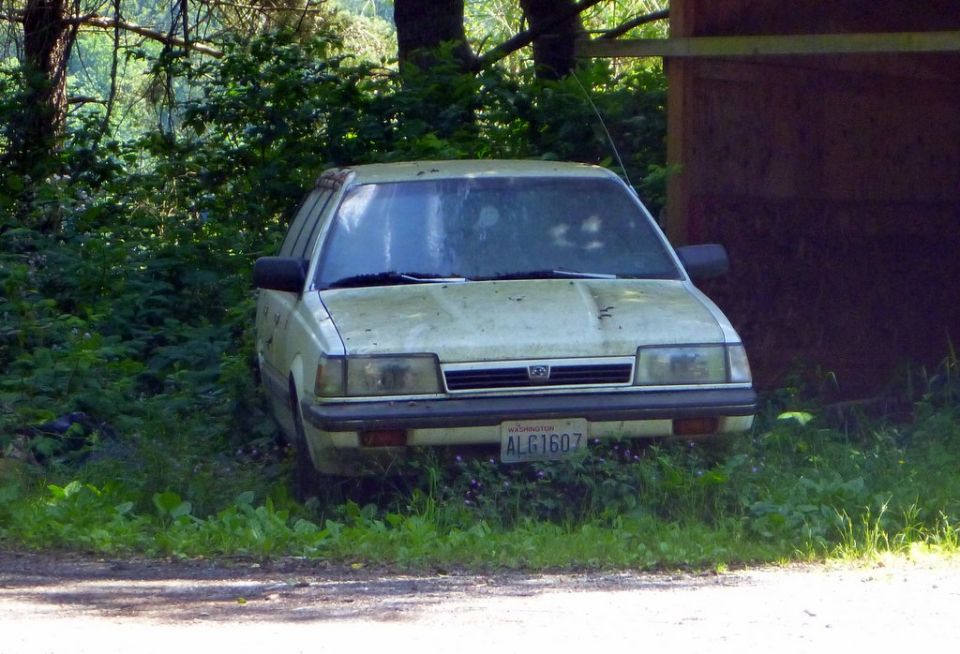 Front view of a dusty, light-colored Subaru sedan parked in overgrown grass next to a wooden structure. License plate reads ALG1607