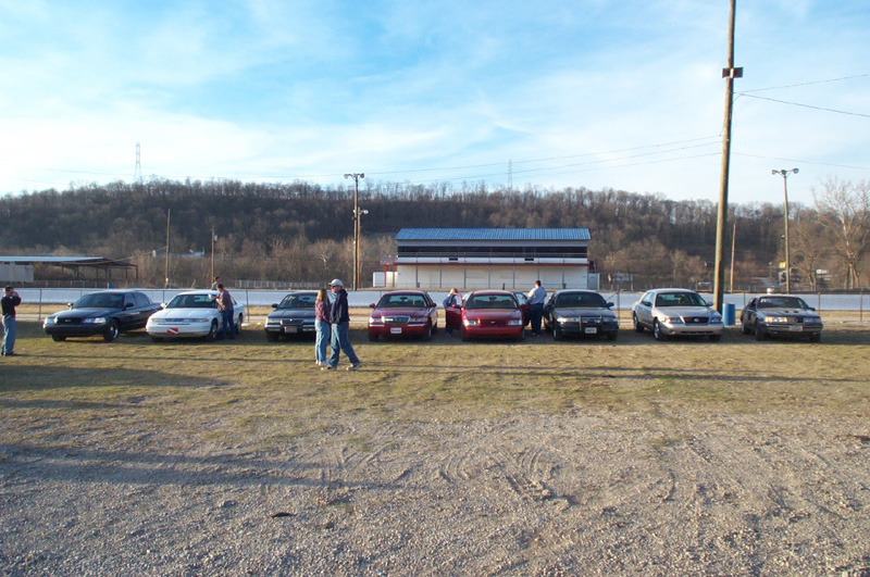 Row of sedans lined up on grass at a track with grandstand and hills in the background
