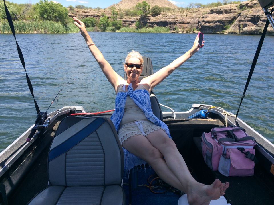 Woman with arms raised on a boat, holding a phone, with a fishing rod and gear visible
