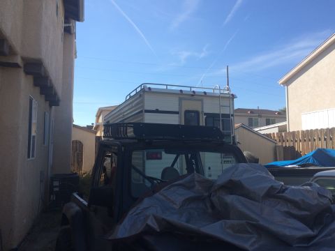1987 Suzuki Samurai - A truck is parked in a residential area, fitted with a camper shell and a roof rack, partially covered by a tarp.