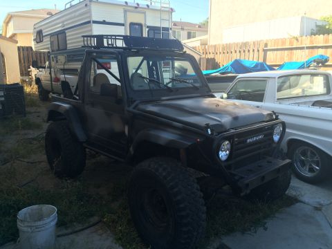 1987 Suzuki Samurai - A modified black 1987 Suzuki Samurai is parked next to a white pickup truck and a camper.