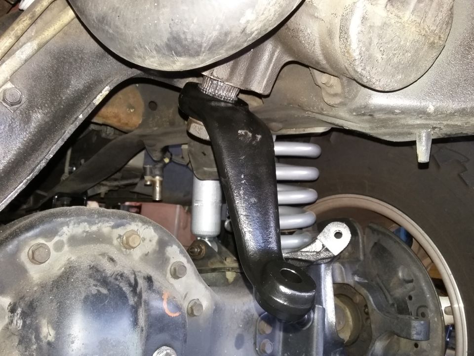 close-up of a vehicle's steering linkage component, possibly an idler arm or pitman arm, with suspension springs and axle visible