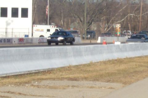 1993 Mercury Grand Marquis - A dark sedan drives down a drag strip with an ambulance and other vehicles in the background.