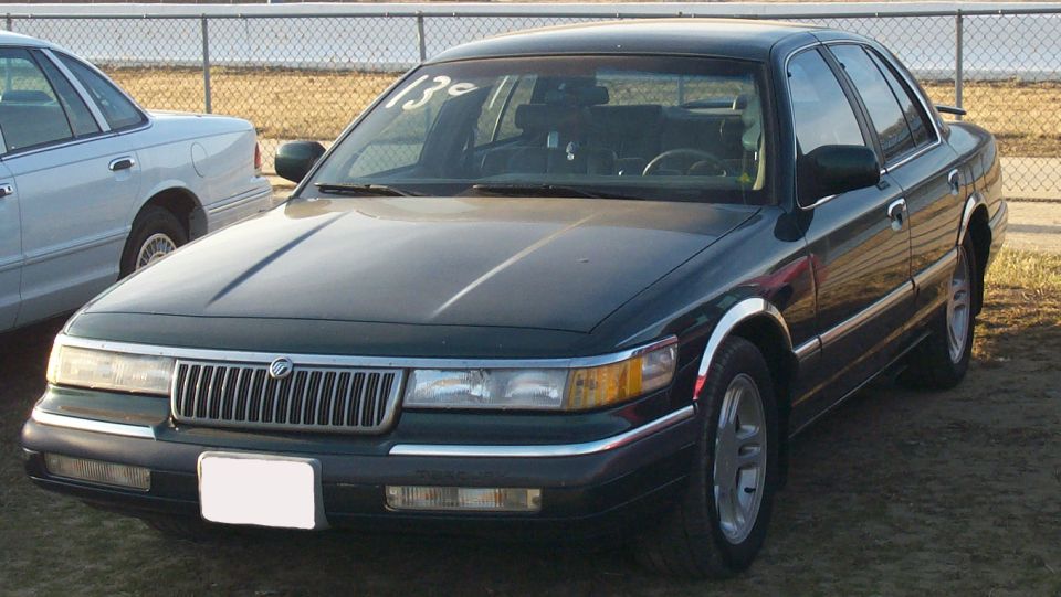 Dark green Mercury Grand Marquis sedan, front three-quarter view, parked by a chain-link fence