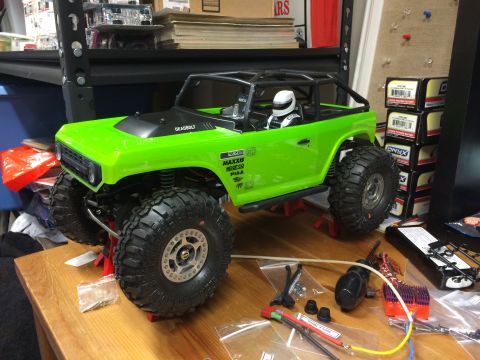 1989 Ford Bronco - A bright green RC car with a driver figure sits on jack stands, surrounded by hobby parts and boxes.