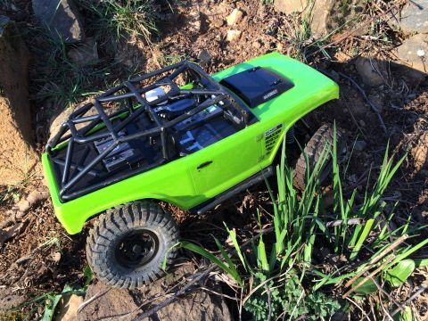 1989 Ford Bronco - This lime green RC rock crawler features an external roll cage and is navigating a rocky, grassy outdoor environment.